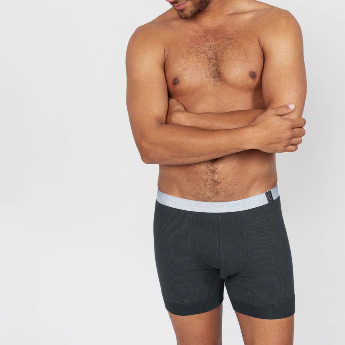 Ein Mann in grauen Boxershorts steht mit verschränkten Armen vor einem hellen Hintergrund.