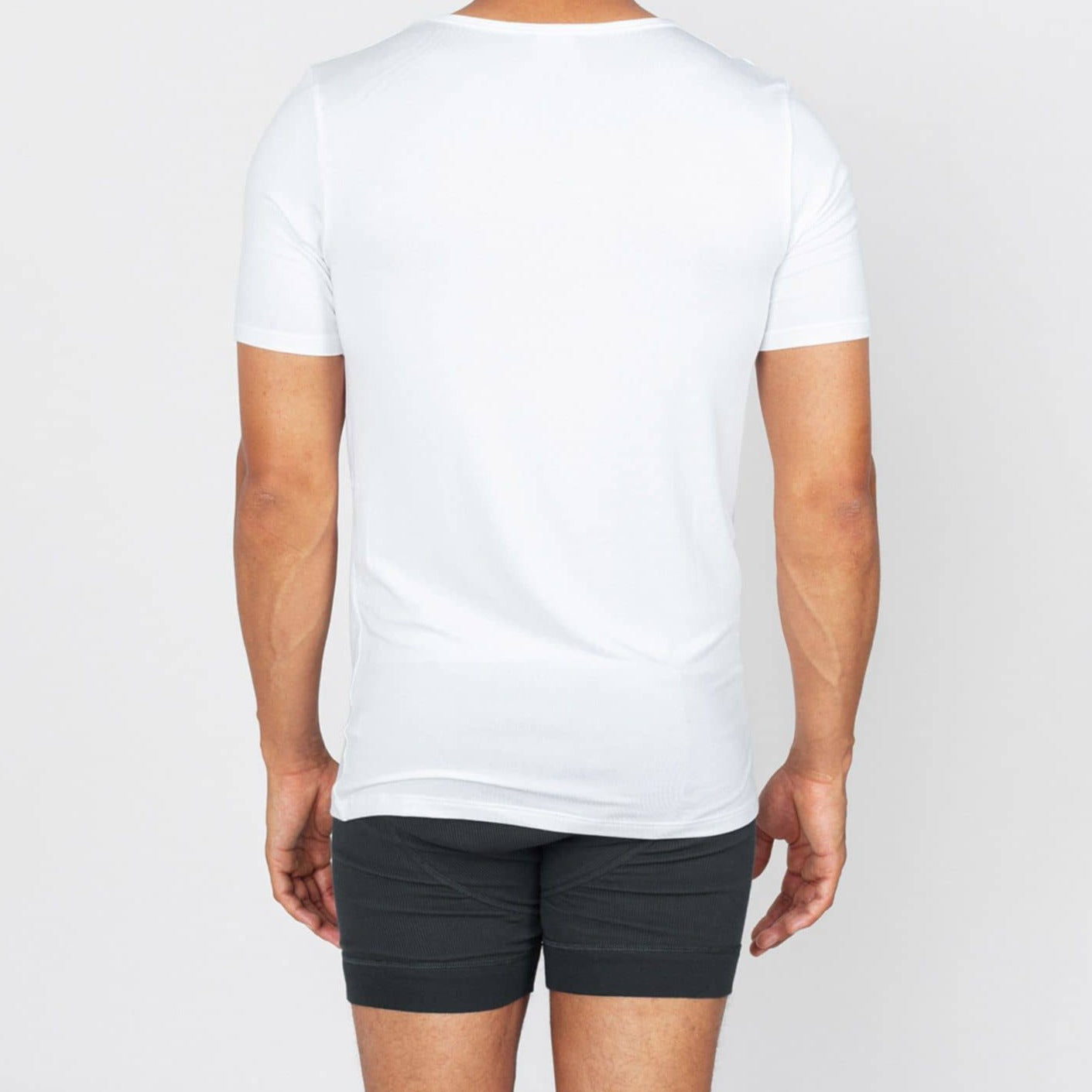Mann von hinten in weißem T-Shirt und schwarzen Shorts, vor einem neutralen Hintergrund stehend.