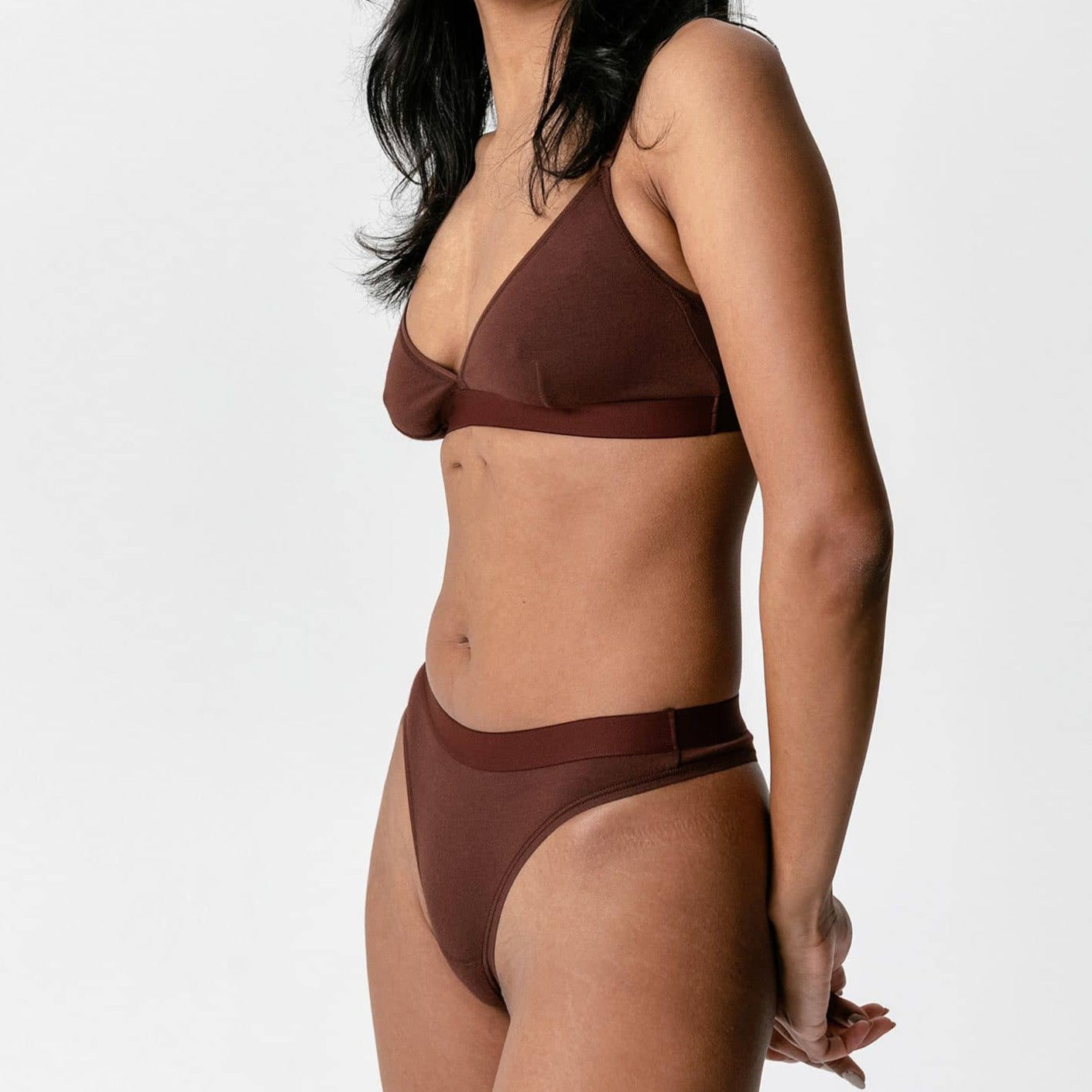 Frau in brauner Unterwäsche steht seitlich vor einem weißen Hintergrund.