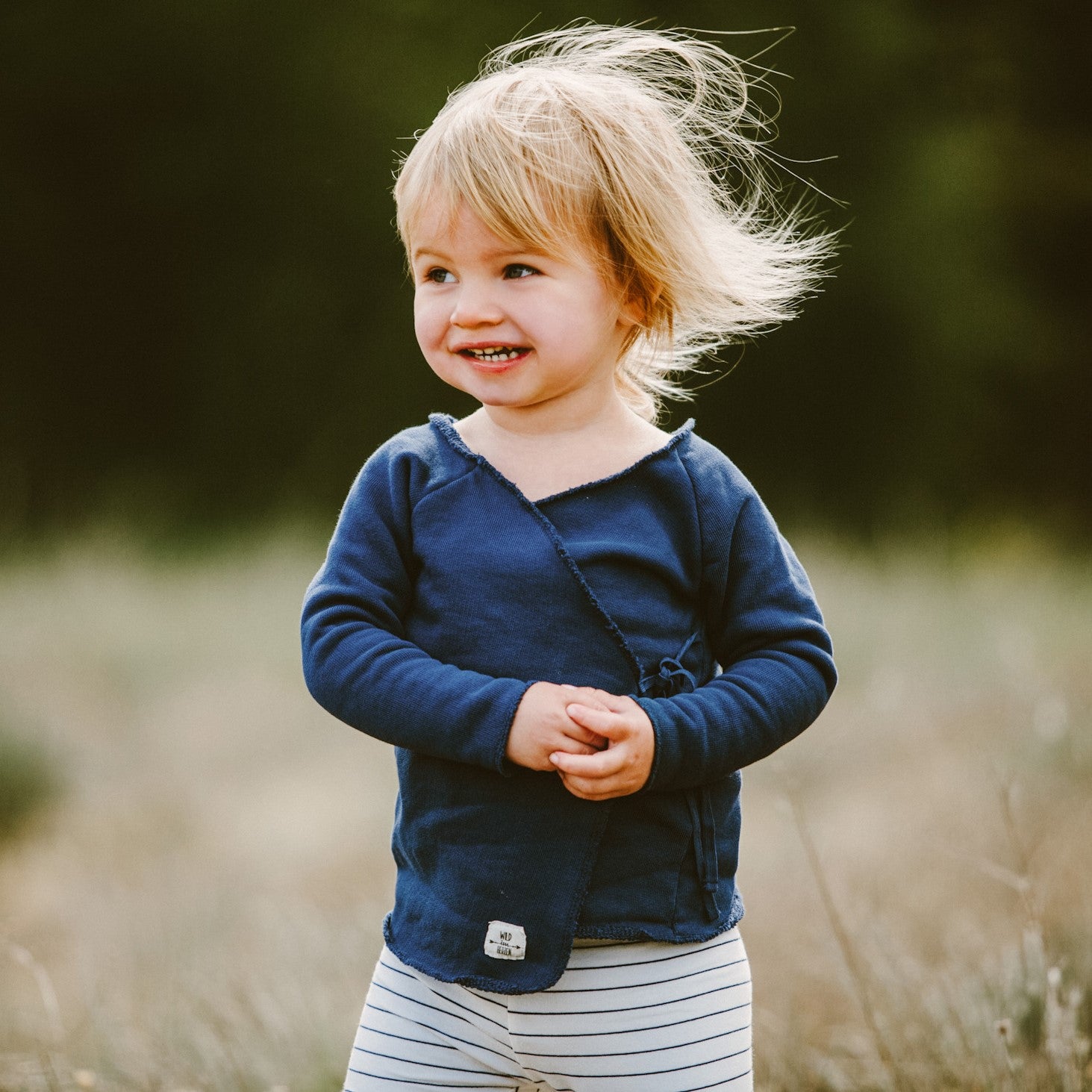 Kleinkind mit blonden Haaren und einem blauen Oberteil lächelt im Freien, während der Wind seine Haare bewegt.