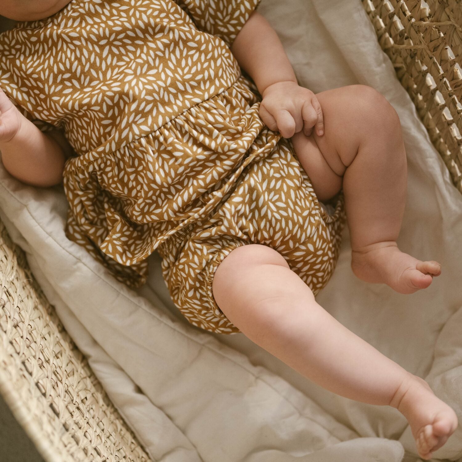 Ein Baby liegt in einem Korb und trägt ein braunes Kleid mit weißem Blattmuster.