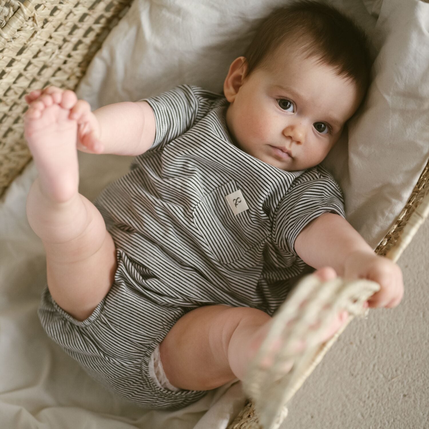 Ein Baby in einem gestreiften Strampler liegt in einem gewebten Korb und greift nach dem Rand.