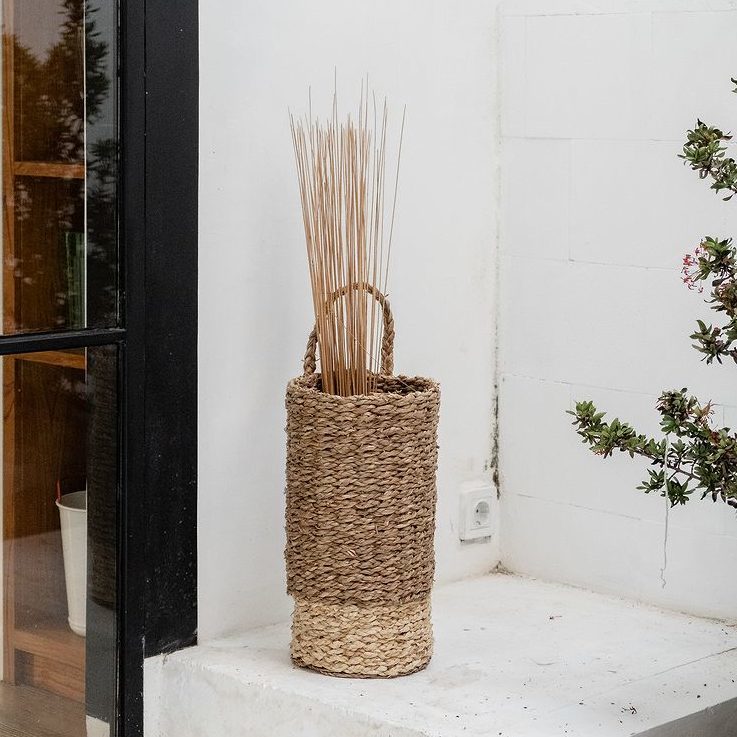 Geflochtener Korb mit Dünengras steht auf weißem Podest neben einer Glastür und einem blühenden Strauch.