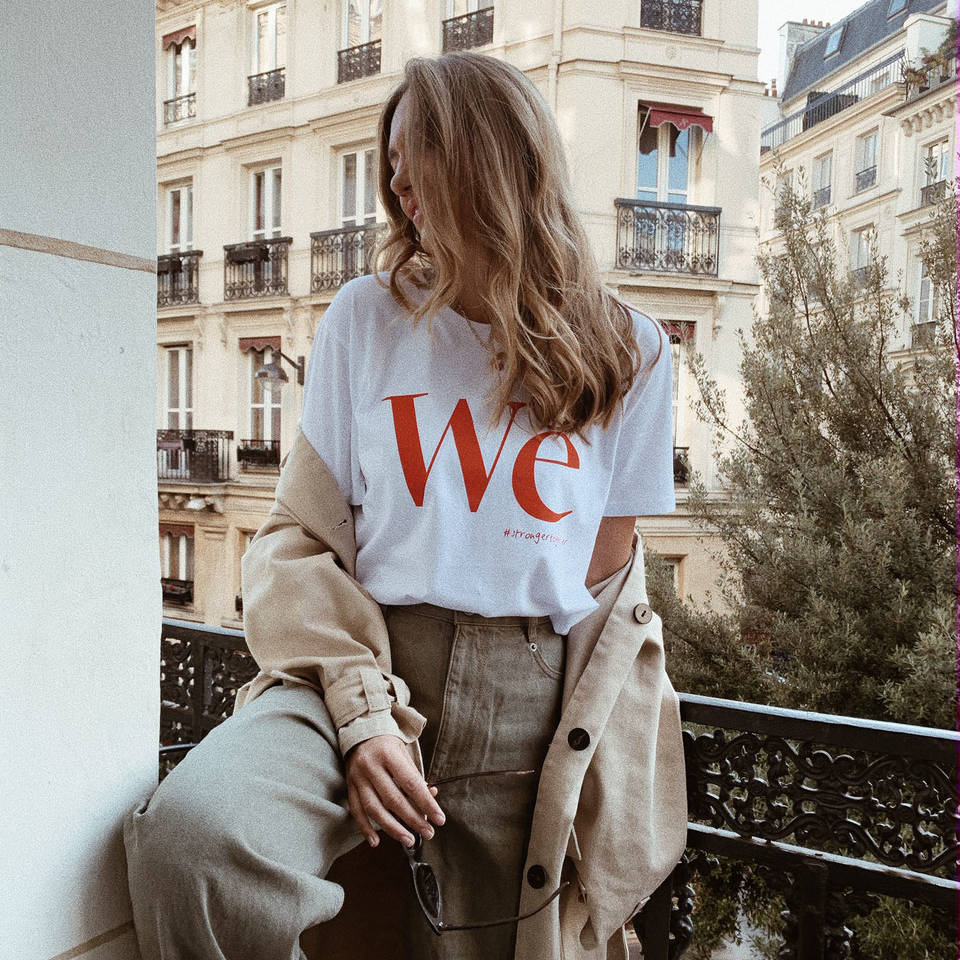 Eine Frau in einem weißen T-Shirt mit "We" darauf, steht auf einem Balkon vor einem Gebäude mit französischen Balkonen.
