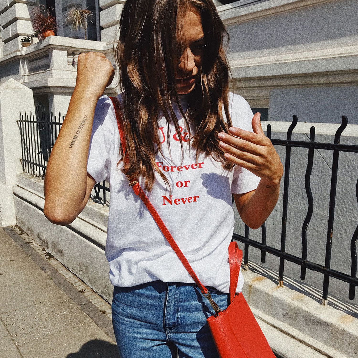 Frau in weißem T-Shirt mit roter Aufschrift, blauer Jeans und roter Tasche steht auf einem Gehweg vor einem Zaun.