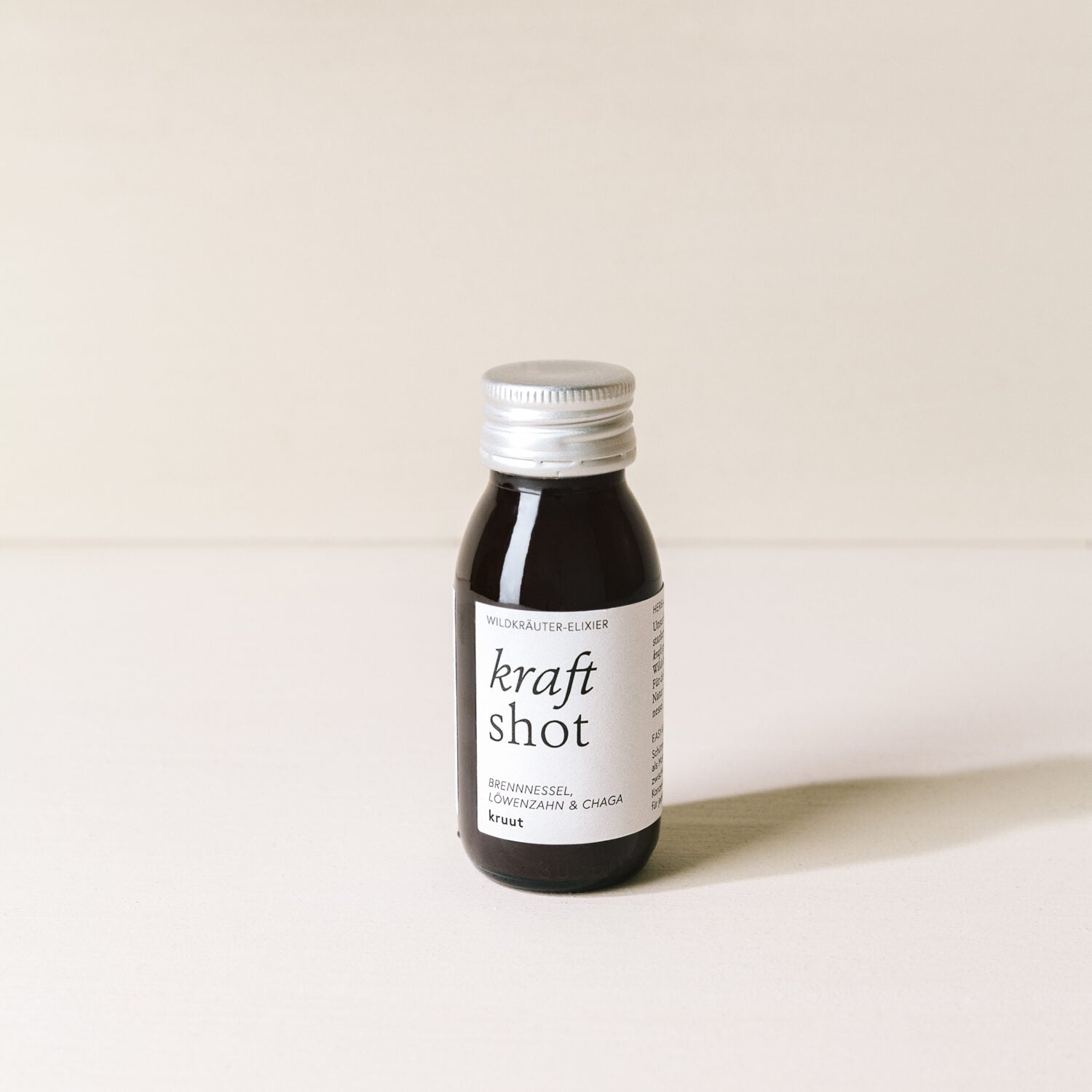 Flasche mit "Kraft Shot" Wildkräuter-Elixier, Brennnessel, Löwenzahn und Chaga, auf hellem Hintergrund.