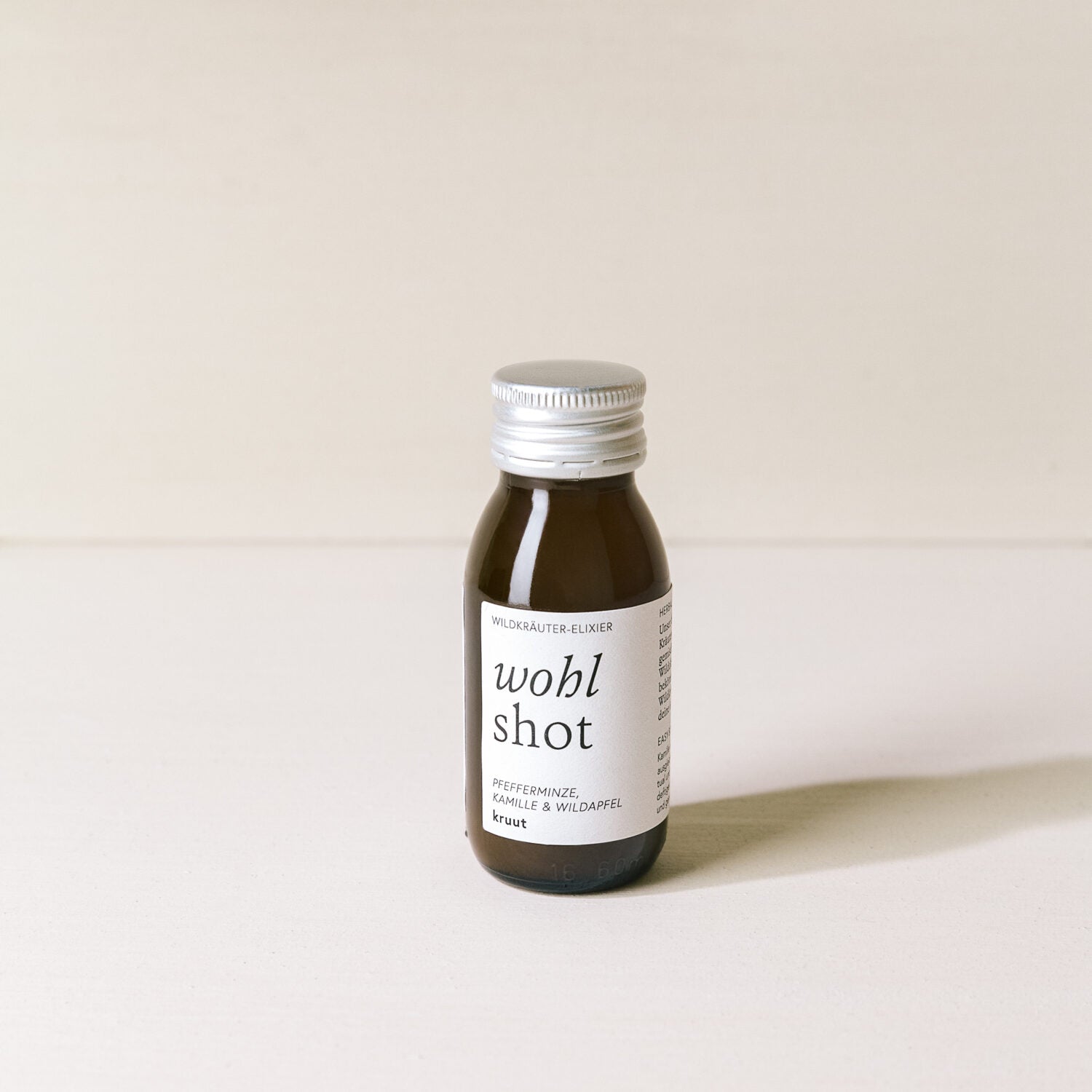 Braune Glasflasche mit der Aufschrift "wohl shot" und Zutaten "Pfefferminze, Kamille & Wildapfel" auf hellem Hintergrund.