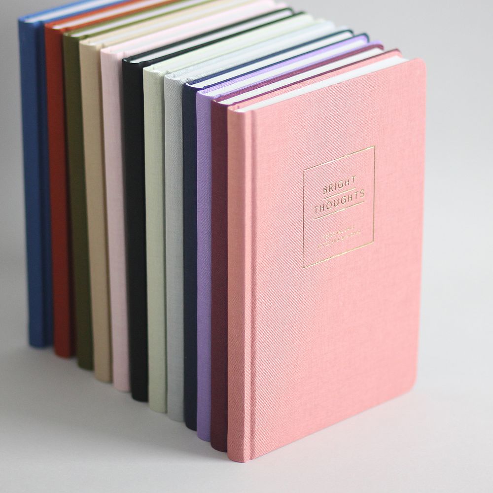 Eine Reihe farbiger Notizbücher steht aufrecht nebeneinander, das vorderste ist rosa und mit "Bright Thoughts" beschriftet.