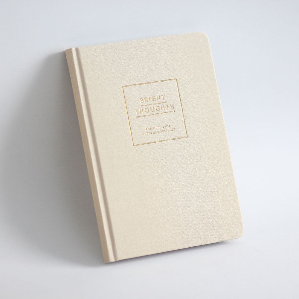 Beiges Notizbuch mit der Aufschrift "Bright Thoughts" auf dem Einband, vor einem hellen Hintergrund.