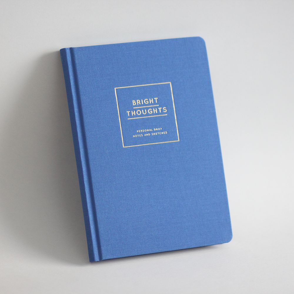 Blaues Notizbuch mit der Aufschrift "Bright Thoughts" in Gold auf dem Einband, vor einem grauen Hintergrund.