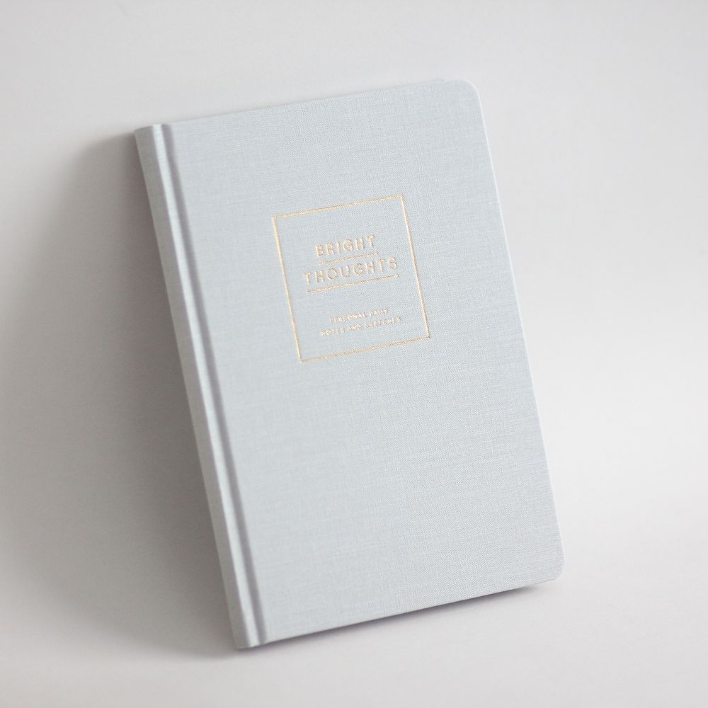 Ein hellgraues Notizbuch mit der Aufschrift "BRIGHT THOUGHTS" in goldener Prägung auf dem Einband.