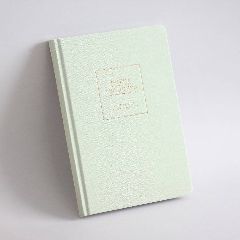 Helles Notizbuch mit der Aufschrift "BRIGHT THOUGHTS" auf dem Einband, auf einem hellgrauen Hintergrund.