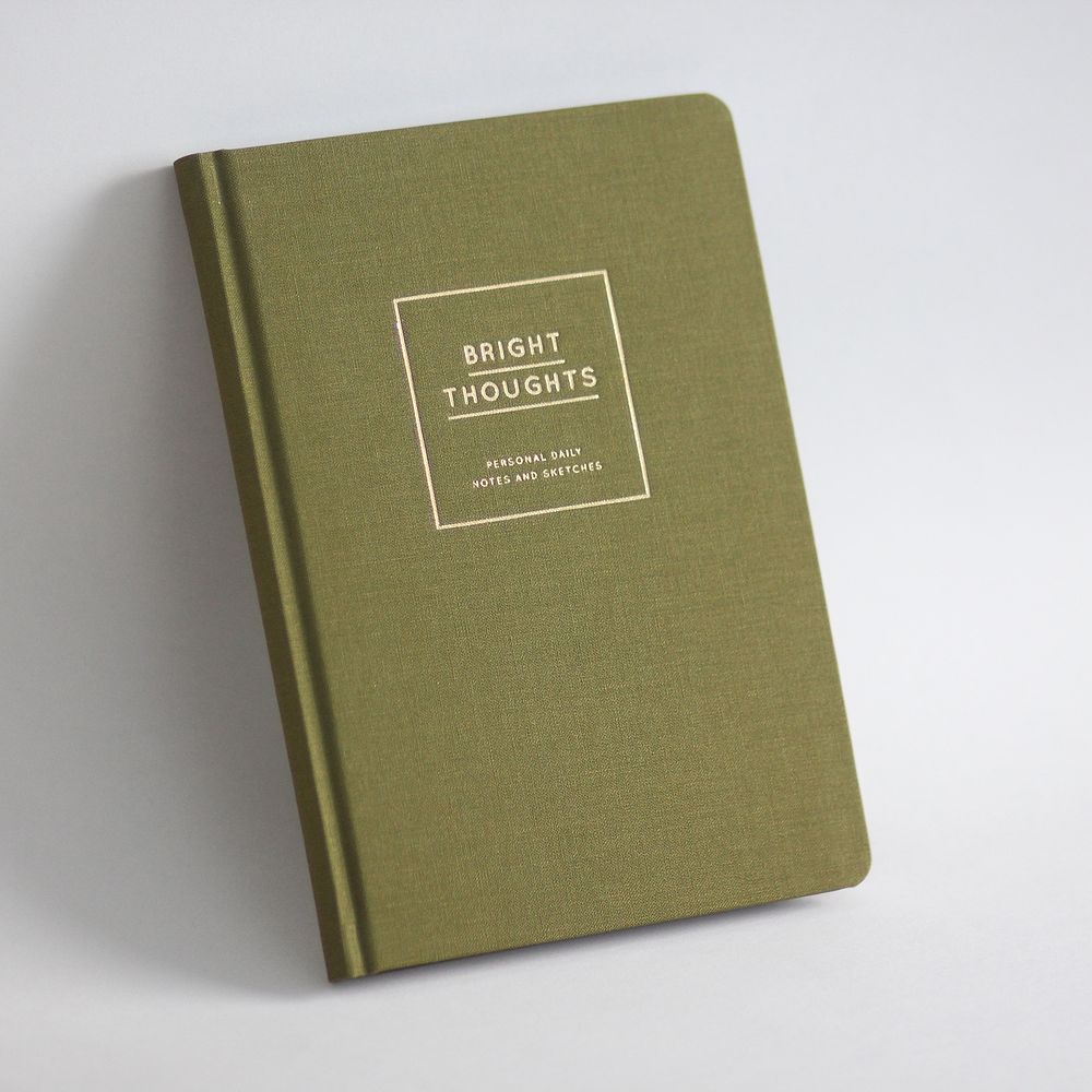 Grünes Notizbuch mit der Aufschrift "Bright Thoughts" auf dem Cover, auf grauem Hintergrund liegend.