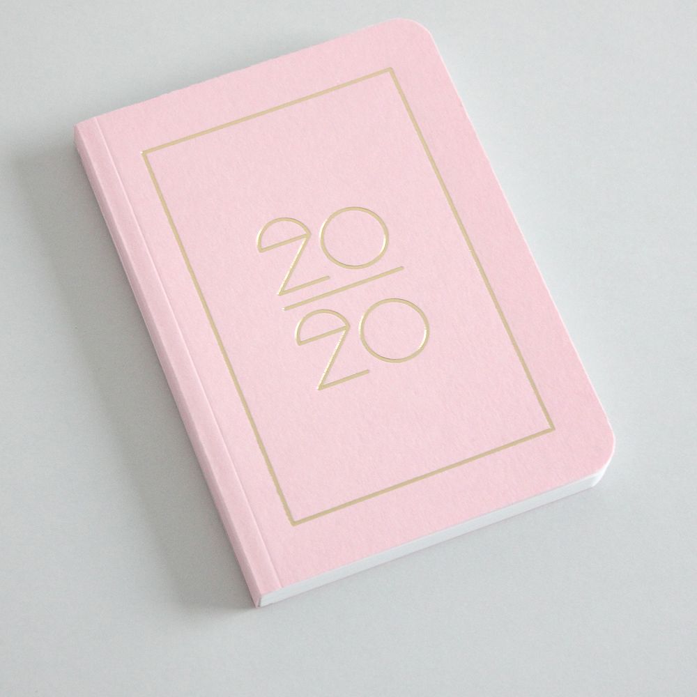 Ein rosafarbenes Notizbuch mit goldener Aufschrift "2020" liegt auf einer grauen Oberfläche.
