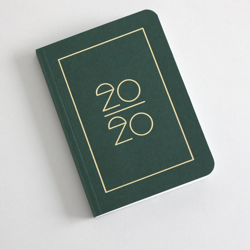 Grünes Notizbuch mit goldener Jahreszahl "2020" auf dem Cover, auf einer hellen Oberfläche liegend.