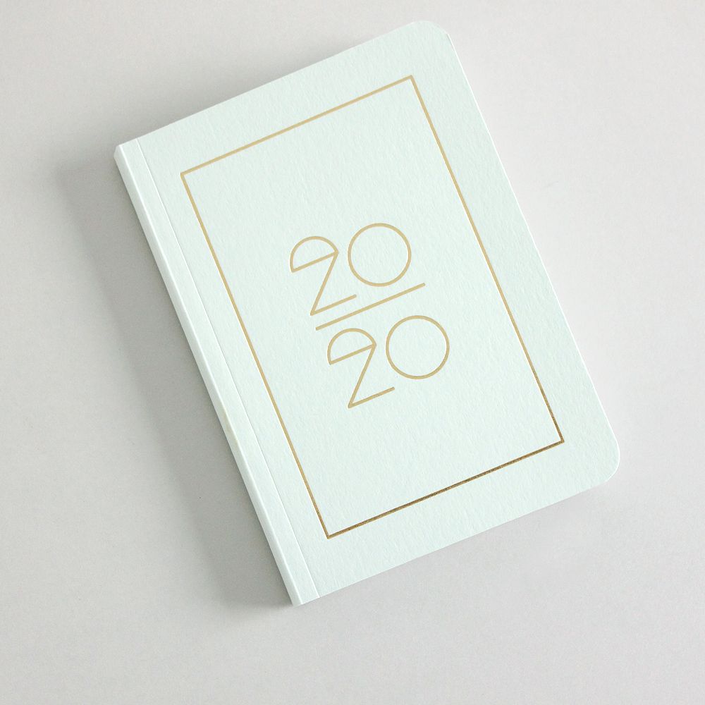 Ein weißes Notizbuch mit der goldenen Aufschrift "20/20" liegt schräg auf einem grauen Hintergrund.