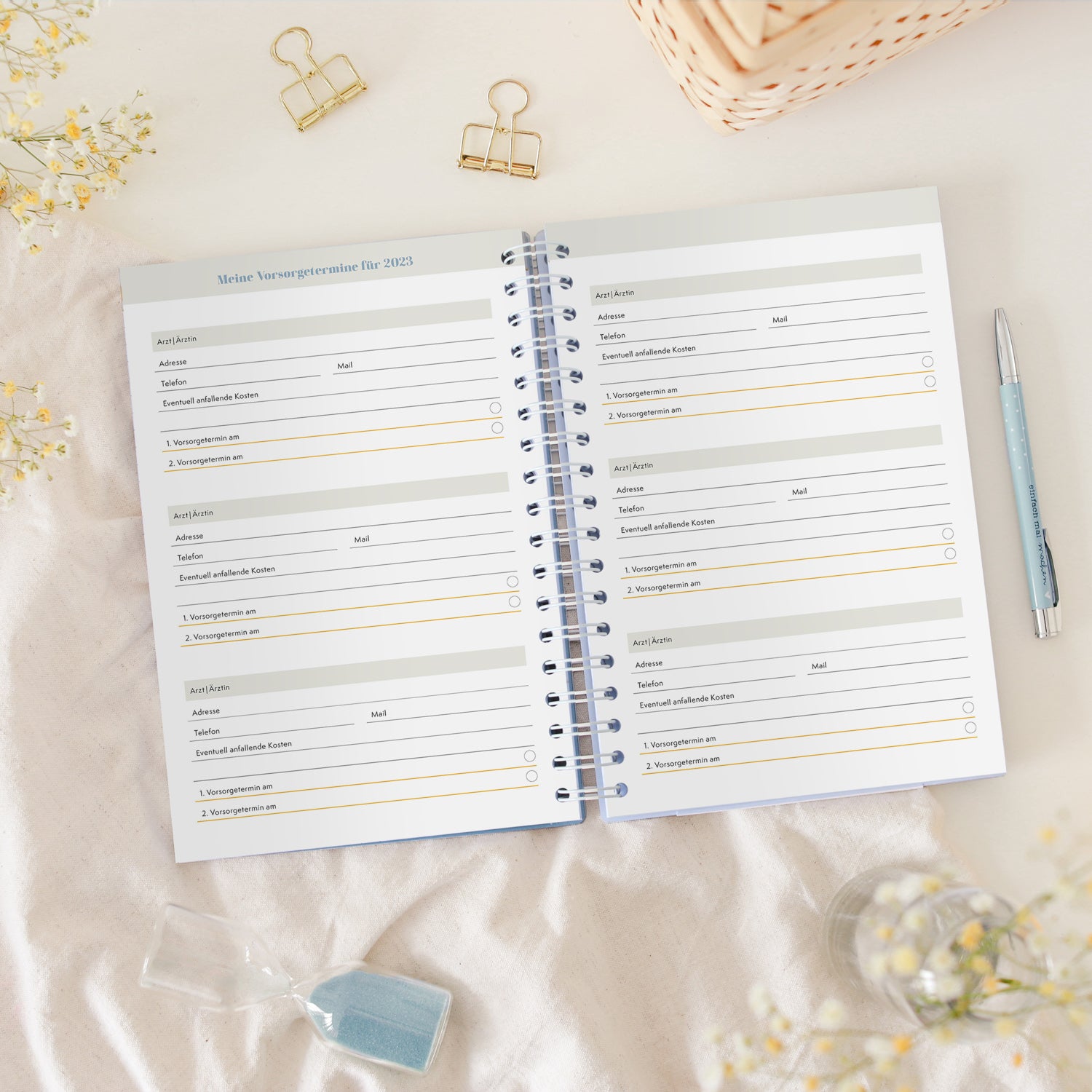 Offener Terminplaner mit Vorsorgeterminen für 2023, umgeben von einem Stift, zwei Klammern und einer Sanduhr.