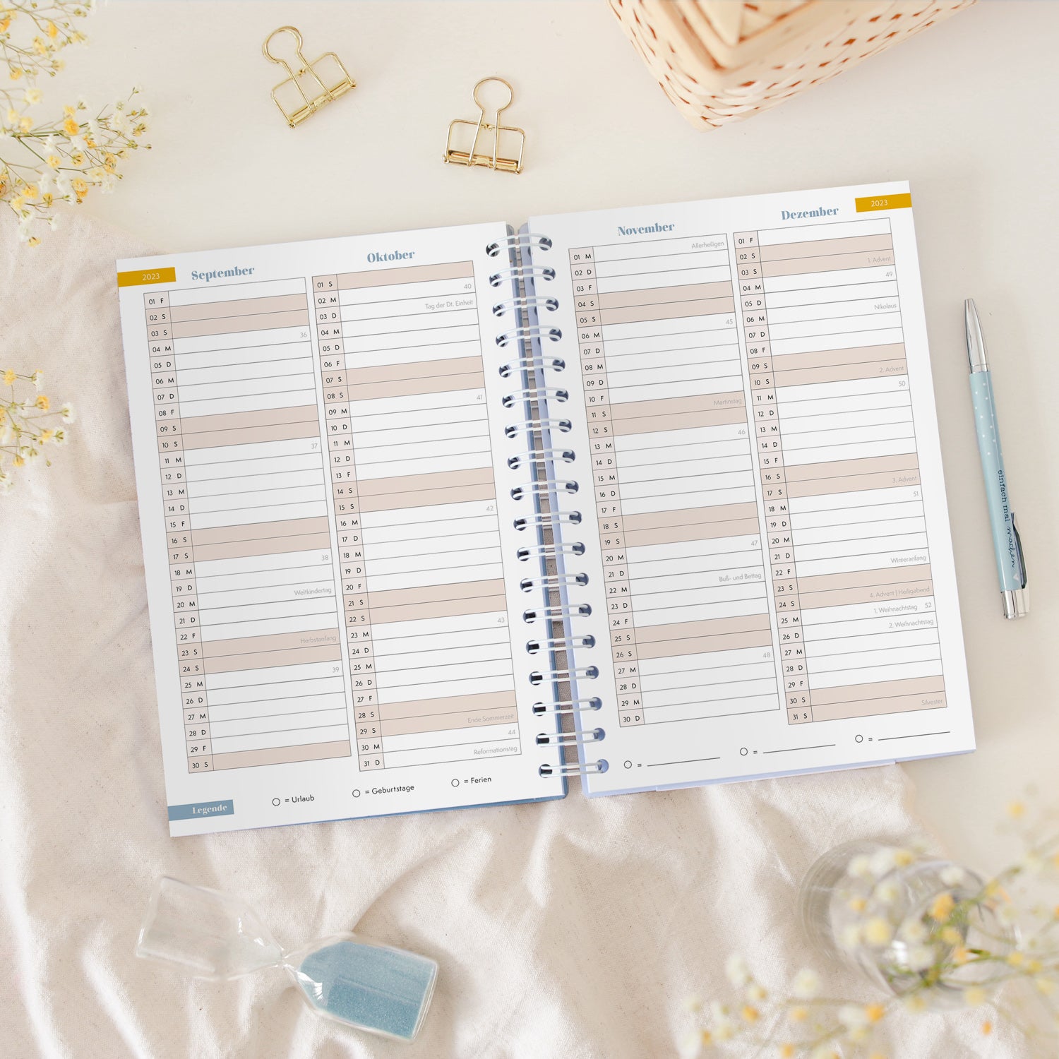Ein aufgeschlagener Planer mit Monatsübersicht, umgeben von zwei goldenen Clips, einem blauen Stift und einer Sanduhr.