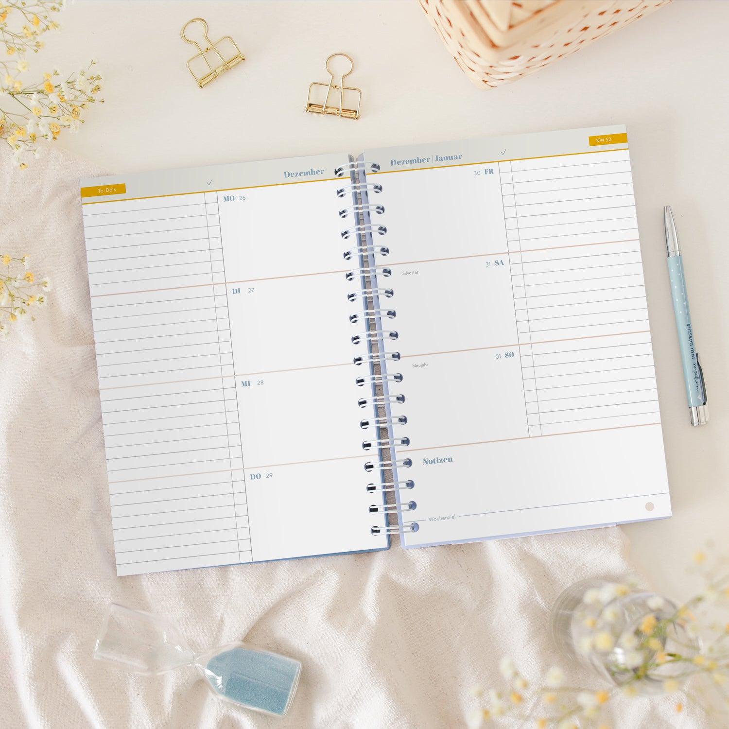 Ein offener Spiralplaner mit Wochenkalender liegt auf einem Tisch, umgeben von Blumen, einem Stift und einer Sanduhr.