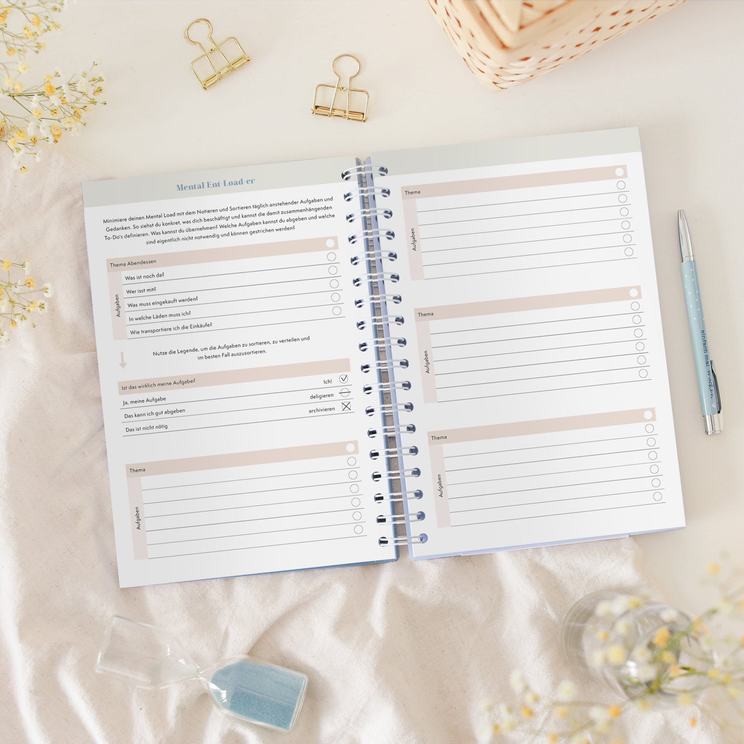 Offenes Notizbuch auf einem Tisch mit zwei goldenen Klammern, einem blauen Kugelschreiber und einem Sanduhr.