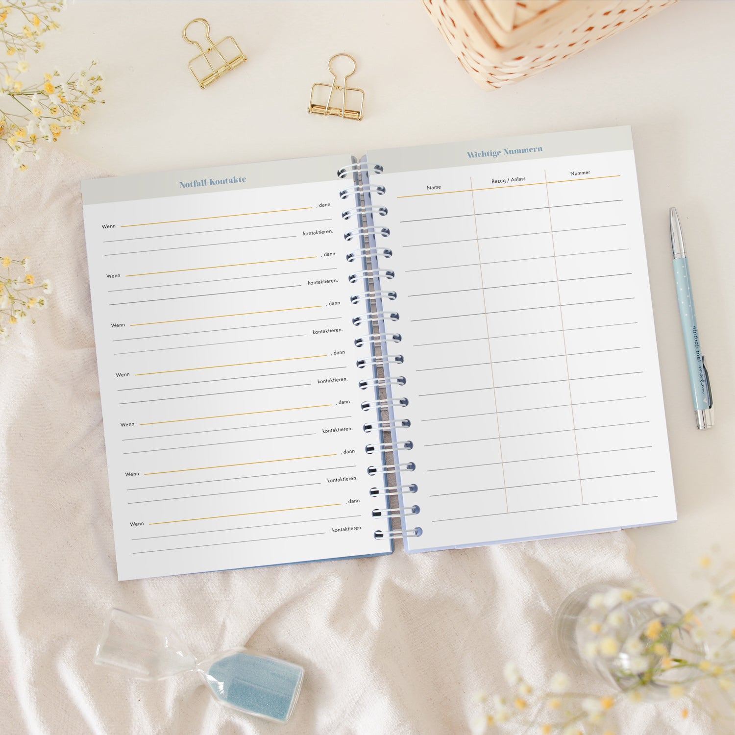 Offenes Notizbuch mit Titeln "Notfallkontakte" und "Wichtige Nummern", umgeben von Büroklammern und einem Stift.