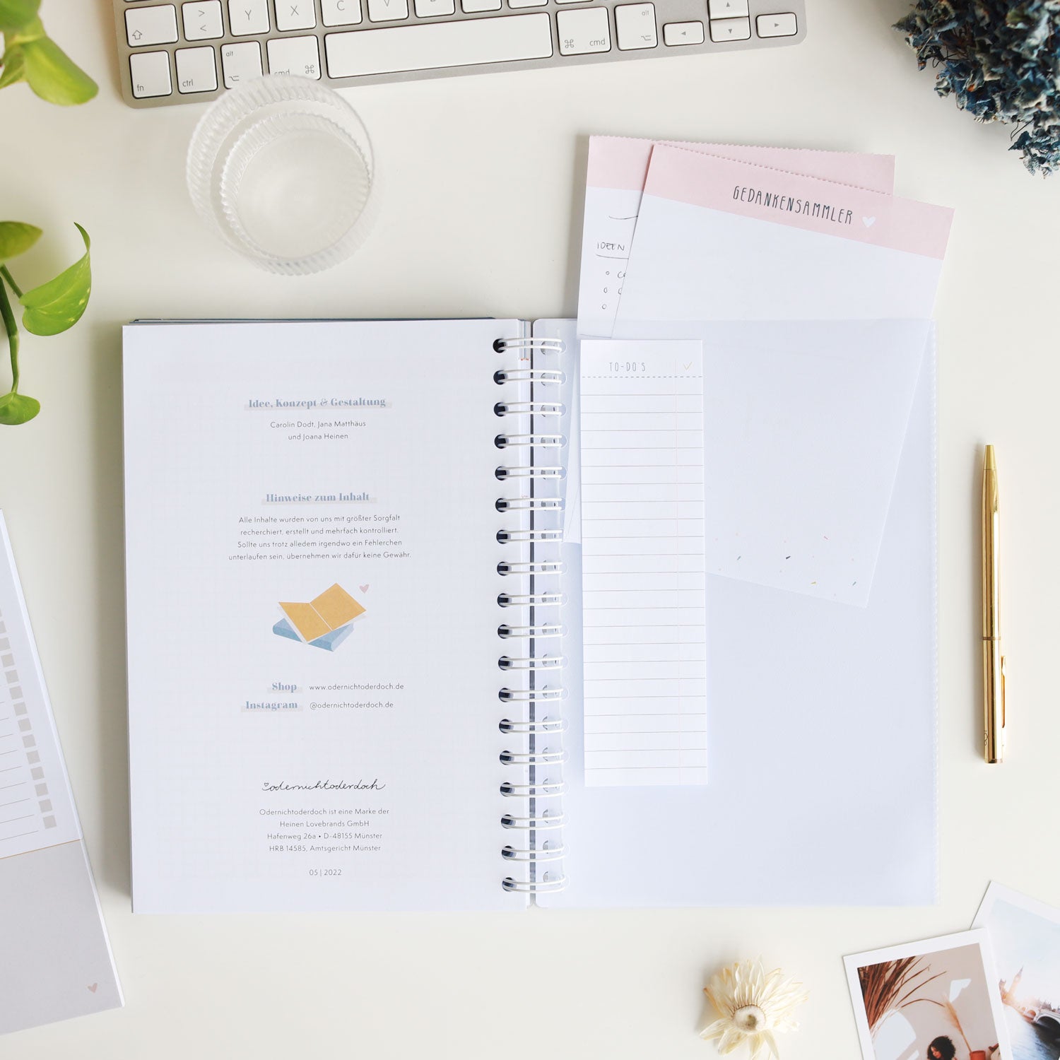 Ein offener Notizblock mit einem Stift, Tastatur, Pflanzenblatt, Fotos und Schreibzubehören auf einem Schreibtisch.