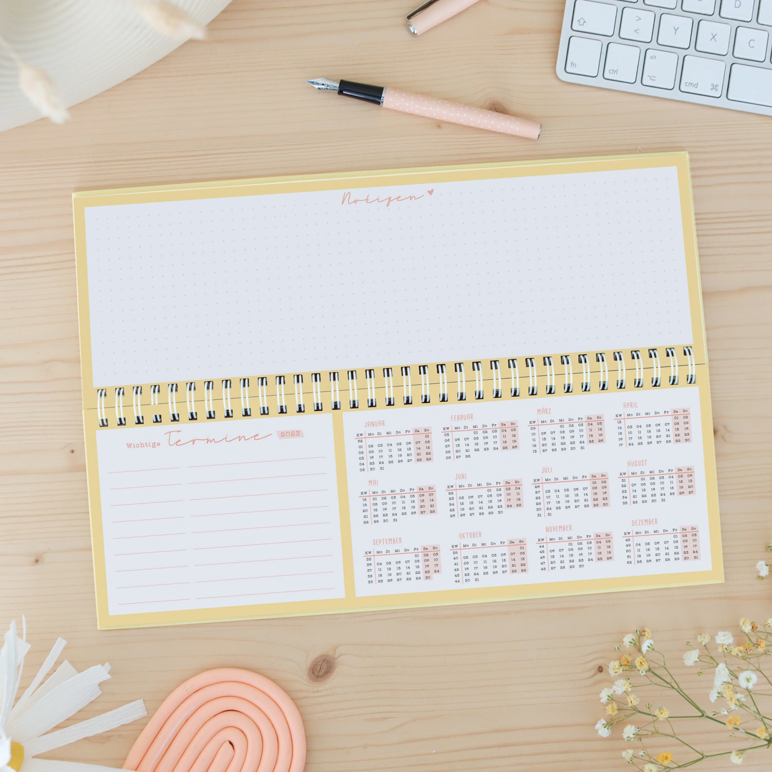 Ein offener Planer mit Kalenderübersicht und Platz für Notizen liegt auf einem Tisch, daneben ein Stift, Blumen und Tastatur.