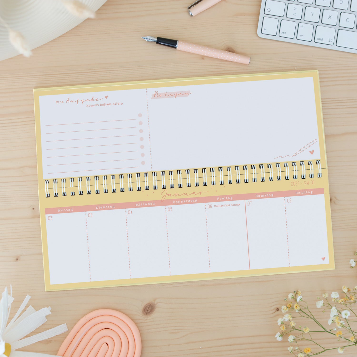 Auf einem Holztisch liegt ein geöffneter Wochenplaner für Januar 2023, daneben ein rosa Stift und eine Tastatur.