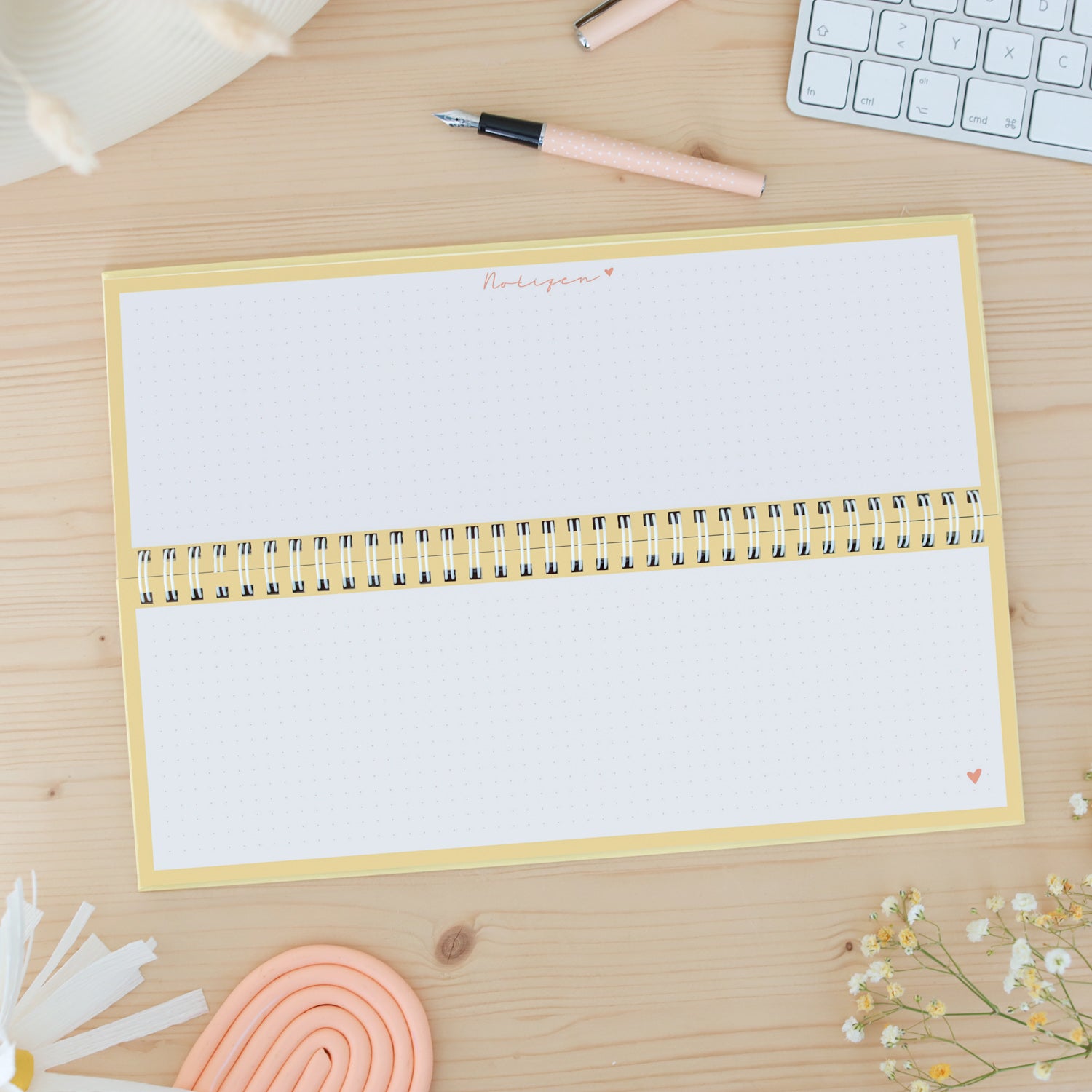Offenes Notizbuch mit Spiralbindung auf einem Holzschreibtisch, umgeben von einem Stift, einer Tastatur und Blumen.