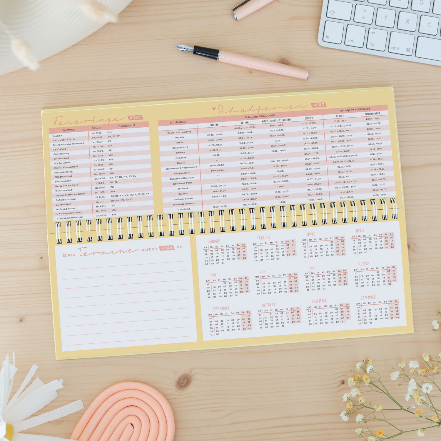 Kalender mit Schulferien und Feiertagen 2023 auf Holzuntergrund, daneben eine Tastatur, ein Stift und Blumen.