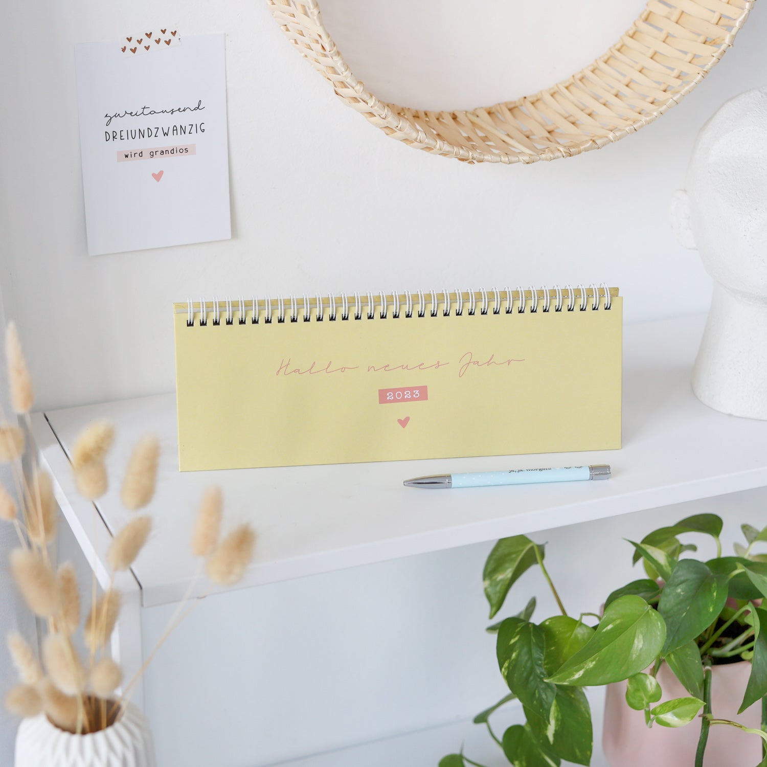 Tischkalender mit der Aufschrift "Hallo neues Jahr 2023" auf einem Regal, daneben eine Pflanze und ein Stift.