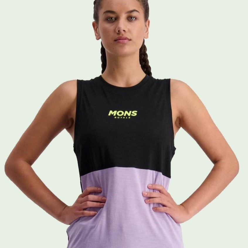 Frau in einem ärmellosen, zweifarbigen Shirt mit der Aufschrift "MONS ROYALE" und verschränkten Armen vor grünem Hintergrund.