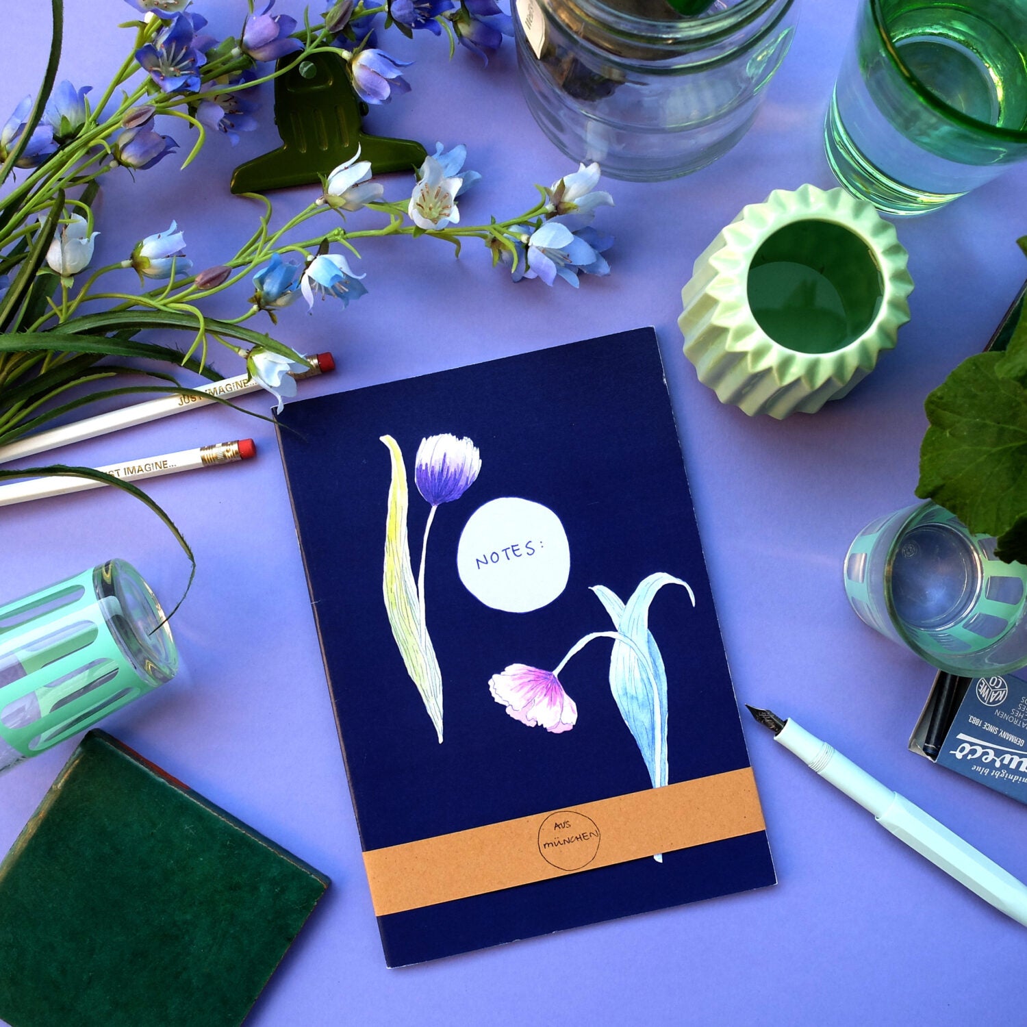 Notizbuch mit Blumenmotiv, umgeben von Stiften, Gläsern und Pflanzen auf einem lila Hintergrund.