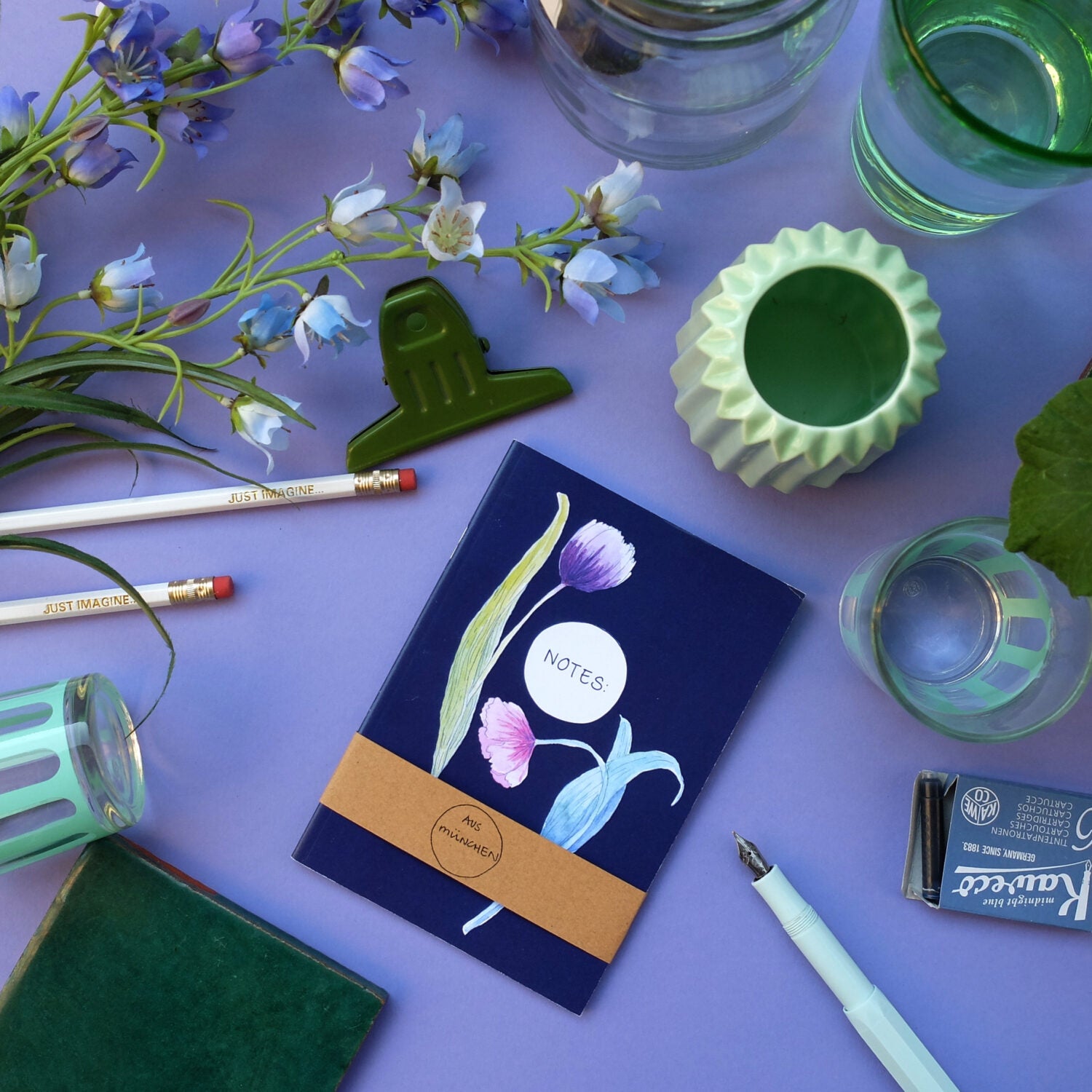 Notizbuch, Stifte, Blüten, grüner Clip und Vasen auf lila Hintergrund arrangiert.