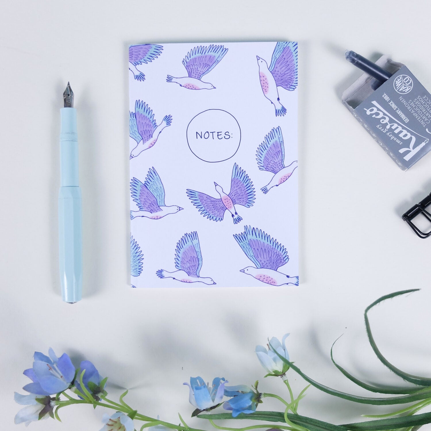 Notizbuch mit fliegenden Vögeln, einem blauen Füller, einem Tintenschachtel, einer Büroklammer und blauen Blumen.