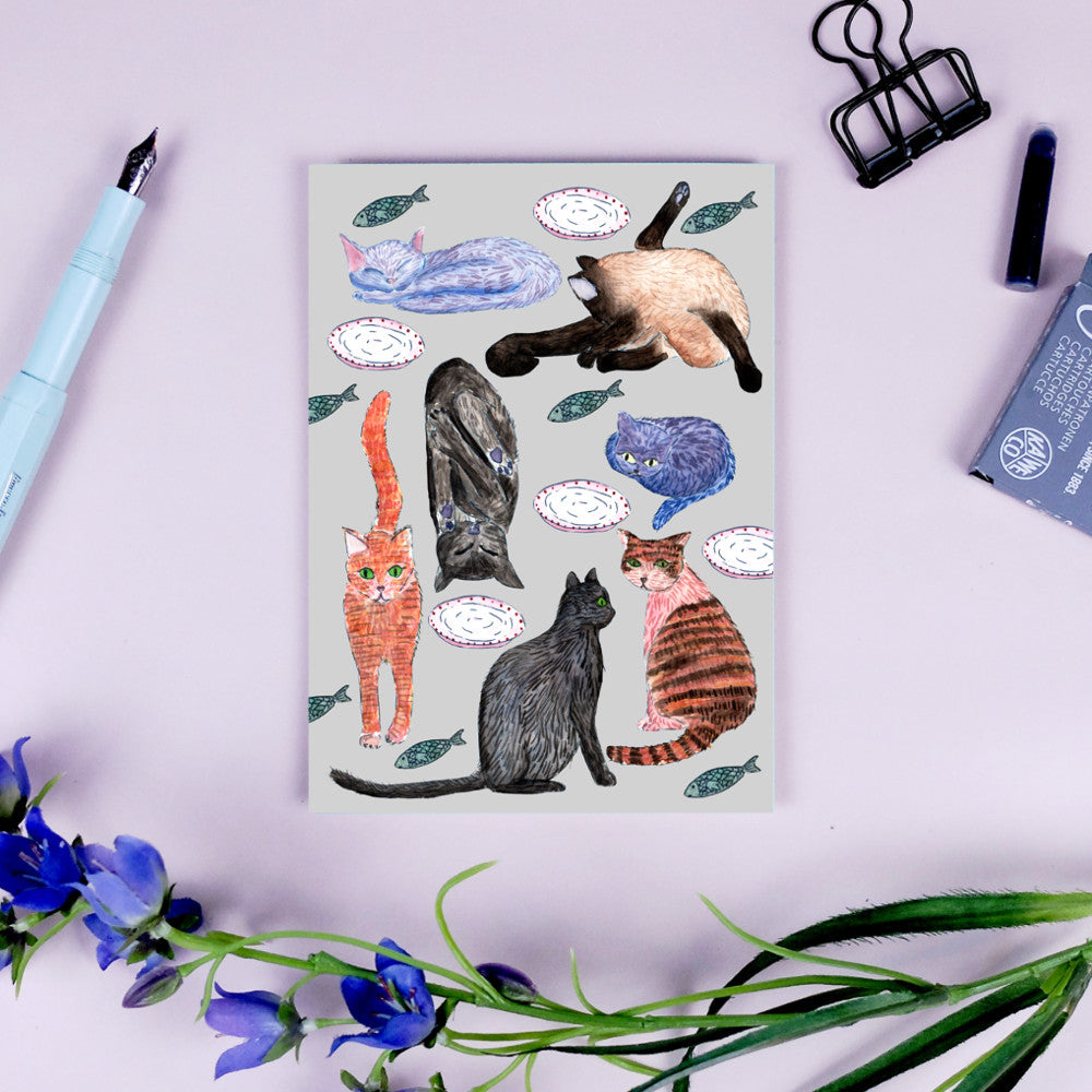 Illustration von Katzen in verschiedenen Positionen auf einer Karte, umgeben von Fischen und Tellern, mit Stiften und Blüten.