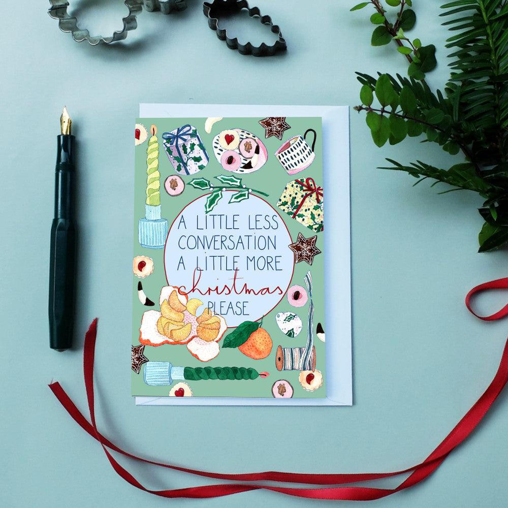 Weihnachtskarte auf einem Tisch mit Tannenzweigen, Backformen, Stift und rotem Band, Text: „A little more Christmas please.”