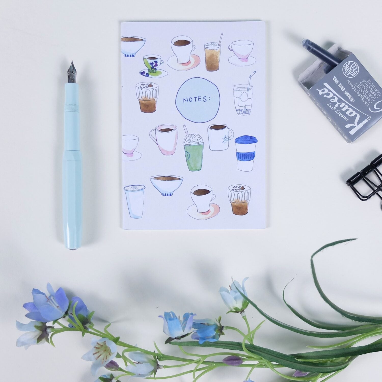 Notizbuch mit gezeichneten Kaffeetassen, blauer Stift, Radiergummi und blaue Blumen auf einem weißen Tisch.