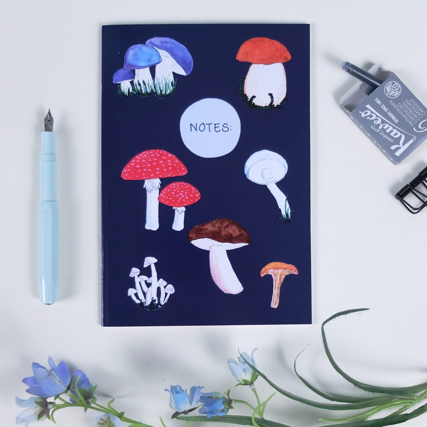 Notizbuch mit Pilzillustrationen, daneben ein blauer Füller, eine Tintenpatrone und ein Blumenstrauß auf weißem Hintergrund.