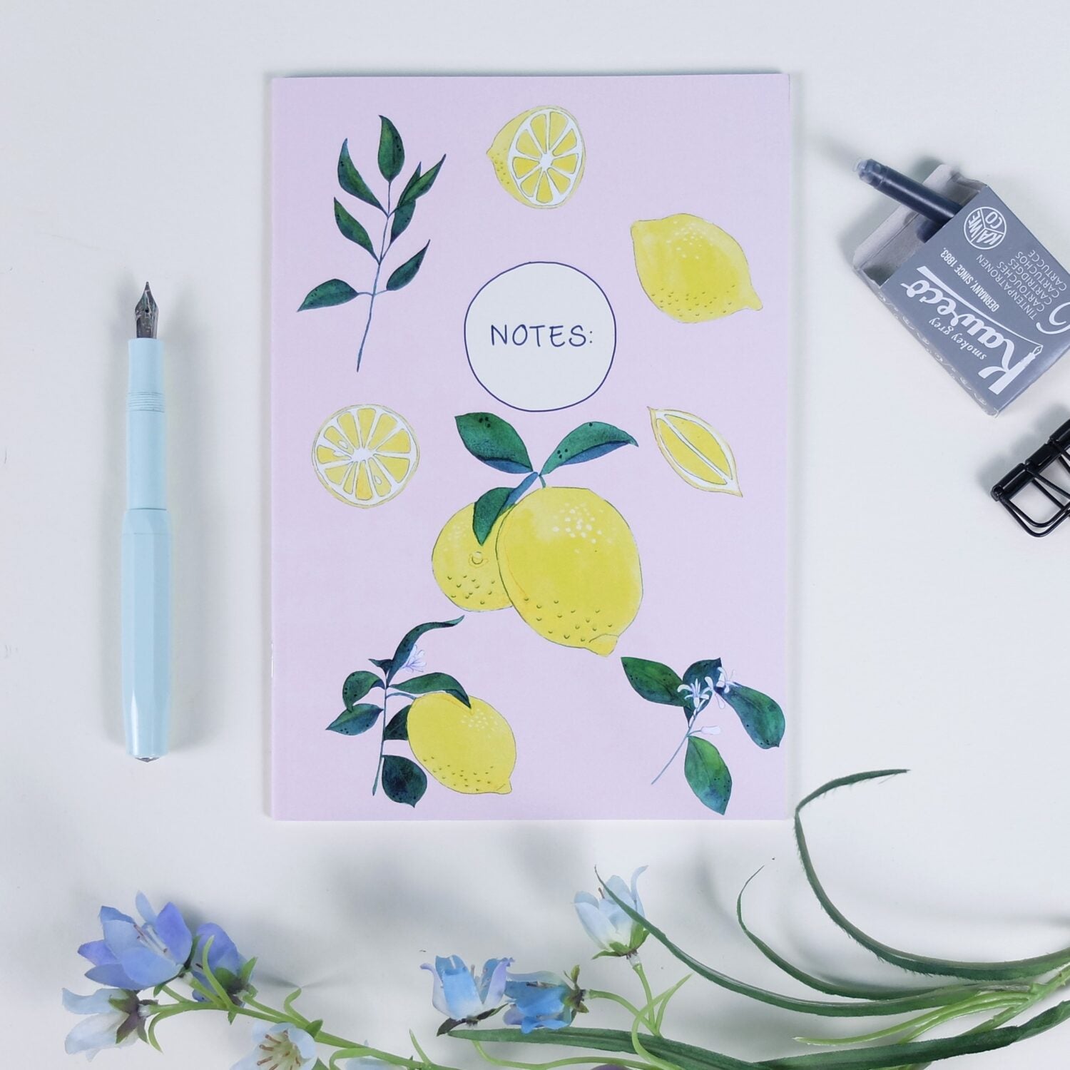 Notizbuch mit Zitronen- und Blattmotiven auf rosa Hintergrund, daneben ein blauer Füller und blaue Blumen.