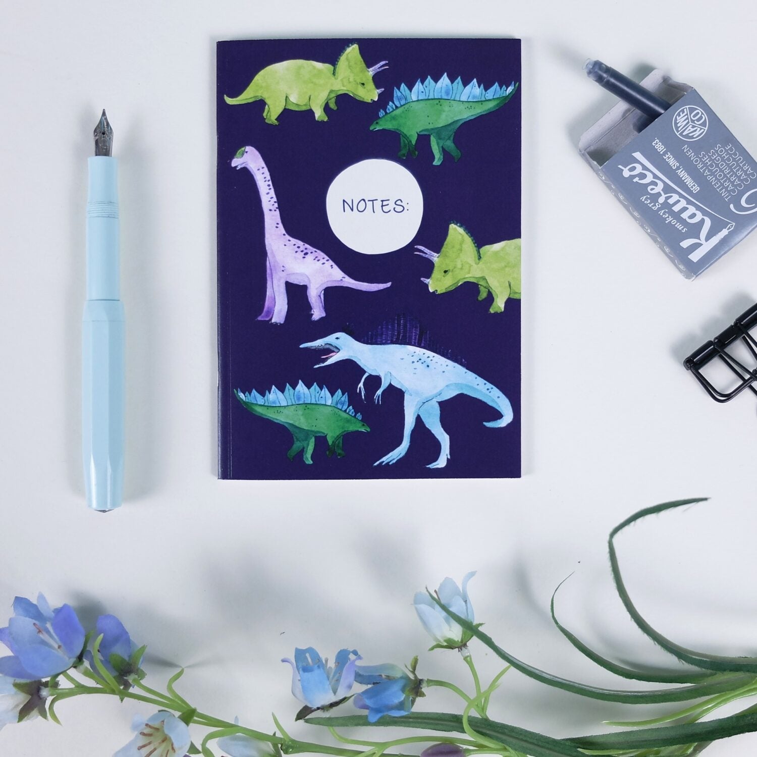 Notizbuch mit Dinosauriermotiven, blauer Füller, Büroklammer, Tintenpatronen und Blumen auf weißem Hintergrund.