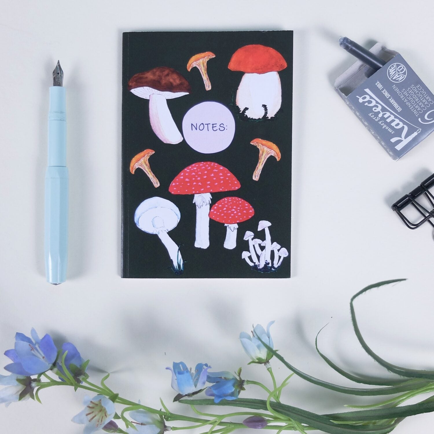 Notizbuch mit Pilzmotiven, daneben ein blauer Füller, Tinte, eine Klammer und blaue Blumen auf weißem Hintergrund.