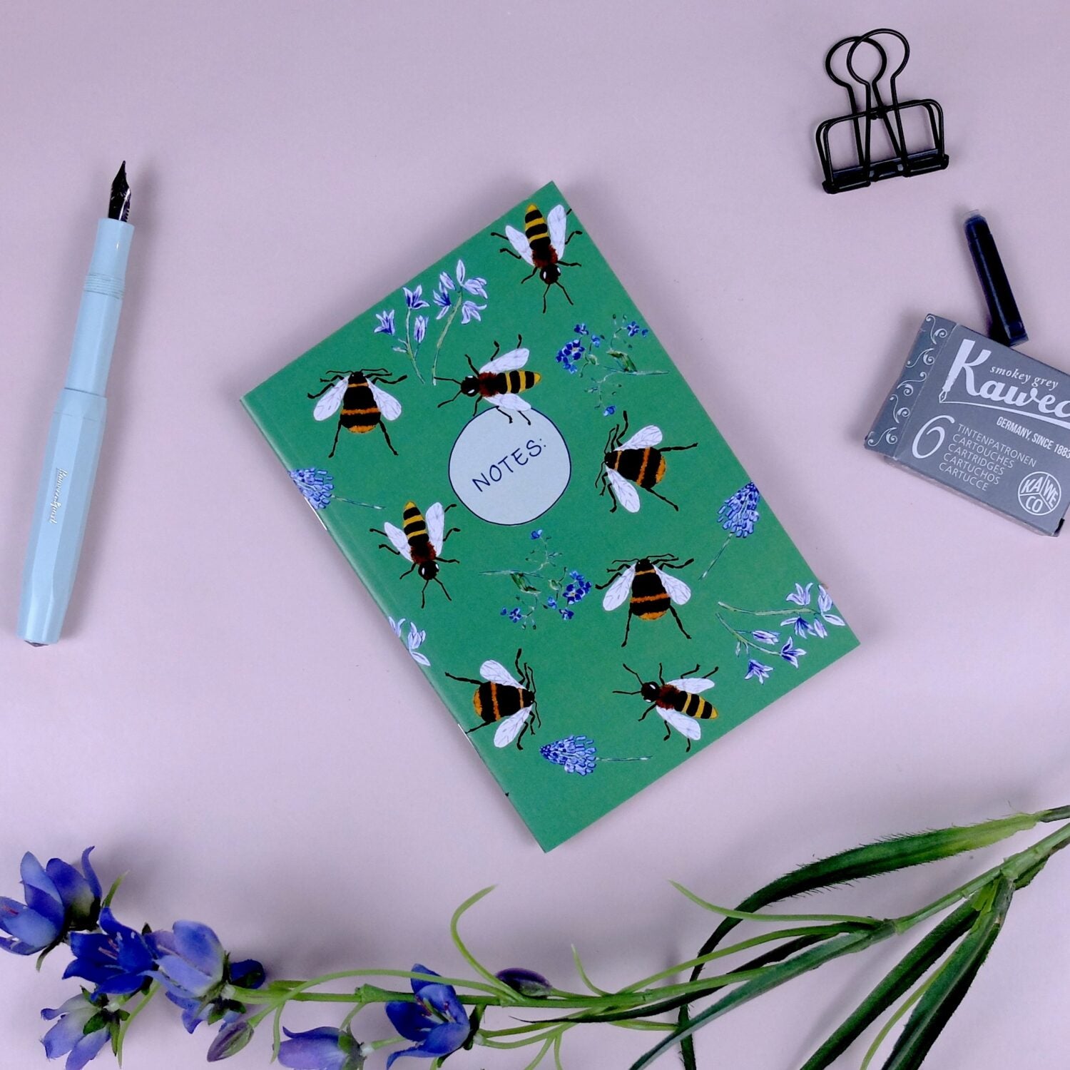 Notizbuch mit Bienenmotiv, hellblauer Füller, Tintenpatronen, Büroklammer und lila Blüten auf rosa Hintergrund.