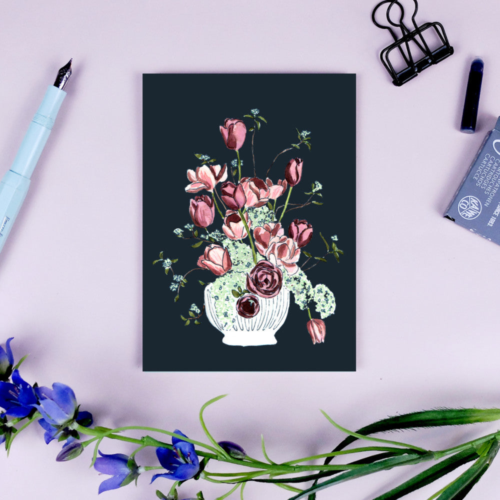 Postkarte mit Blumenvase-Illustration auf dunklem Hintergrund, umgeben von Stift und Klammern auf rosa Oberfläche.