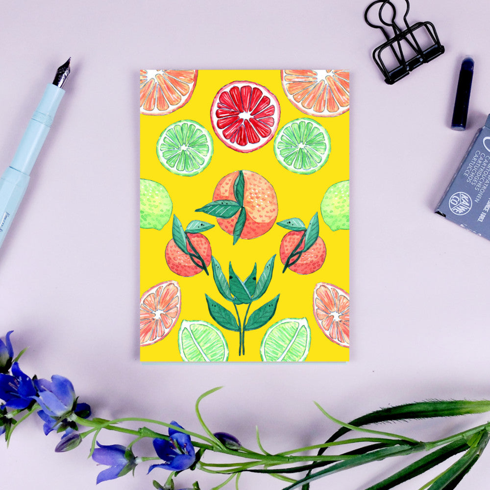 Bunte Illustration von Zitrusfrüchten auf gelbem Hintergrund, umrahmt von einem Füller, Büroklammer und Blumen.