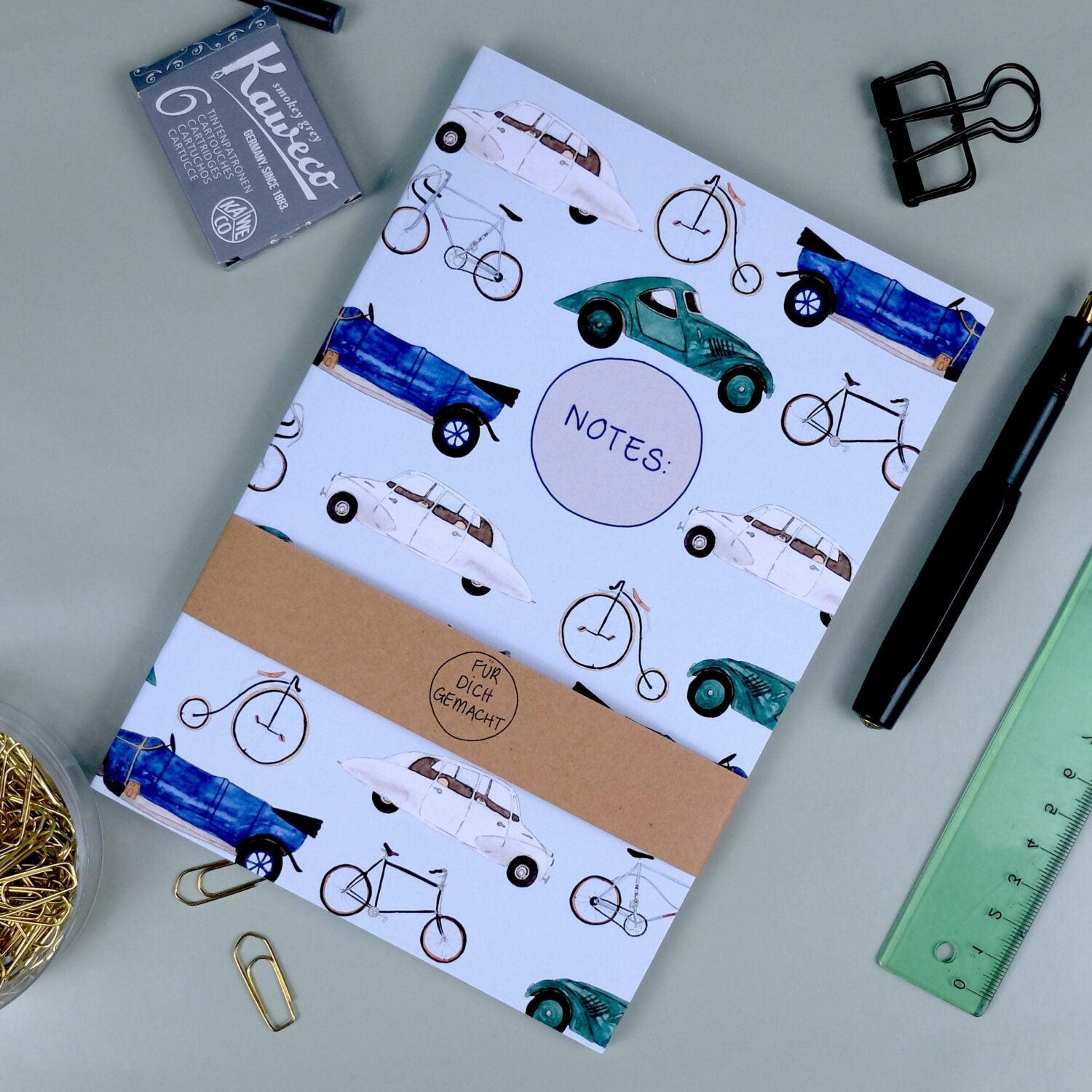 Notizbuch mit Auto- und Fahrradmuster, umgeben von Büroklammern, Bleistift, Lineal und schwarzem Binderclip.