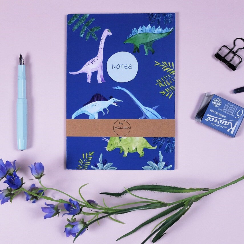 Notizheft mit Dinosaurierdesign auf blauem Hintergrund, mit Kuli, Radiergummi, Büroklammer und Blumenarrangement.