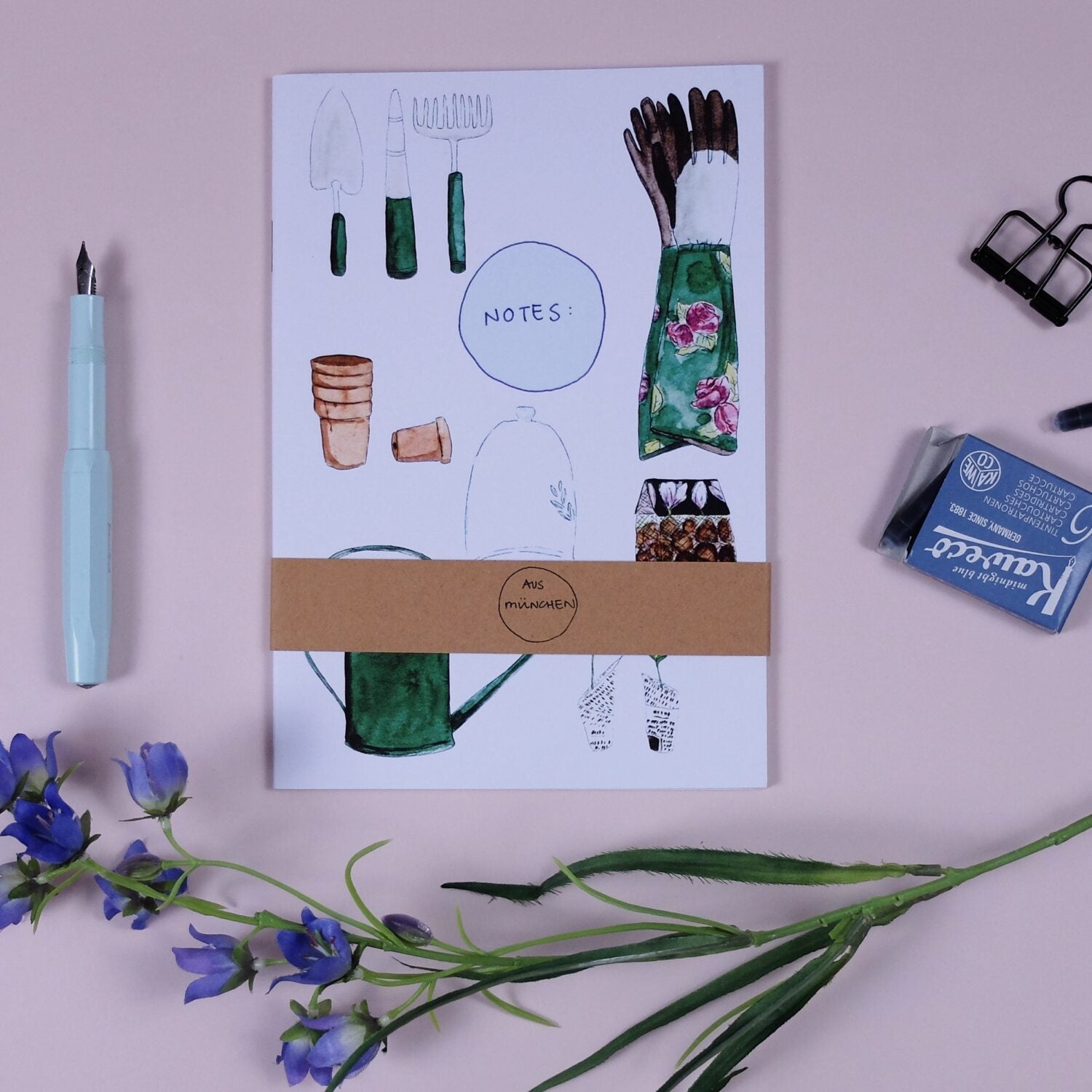 Notizbuch mit Gartenzubehör-Illustrationen, blauer Stift, Radiergummi und blaue Blumen auf einem rosa Hintergrund.