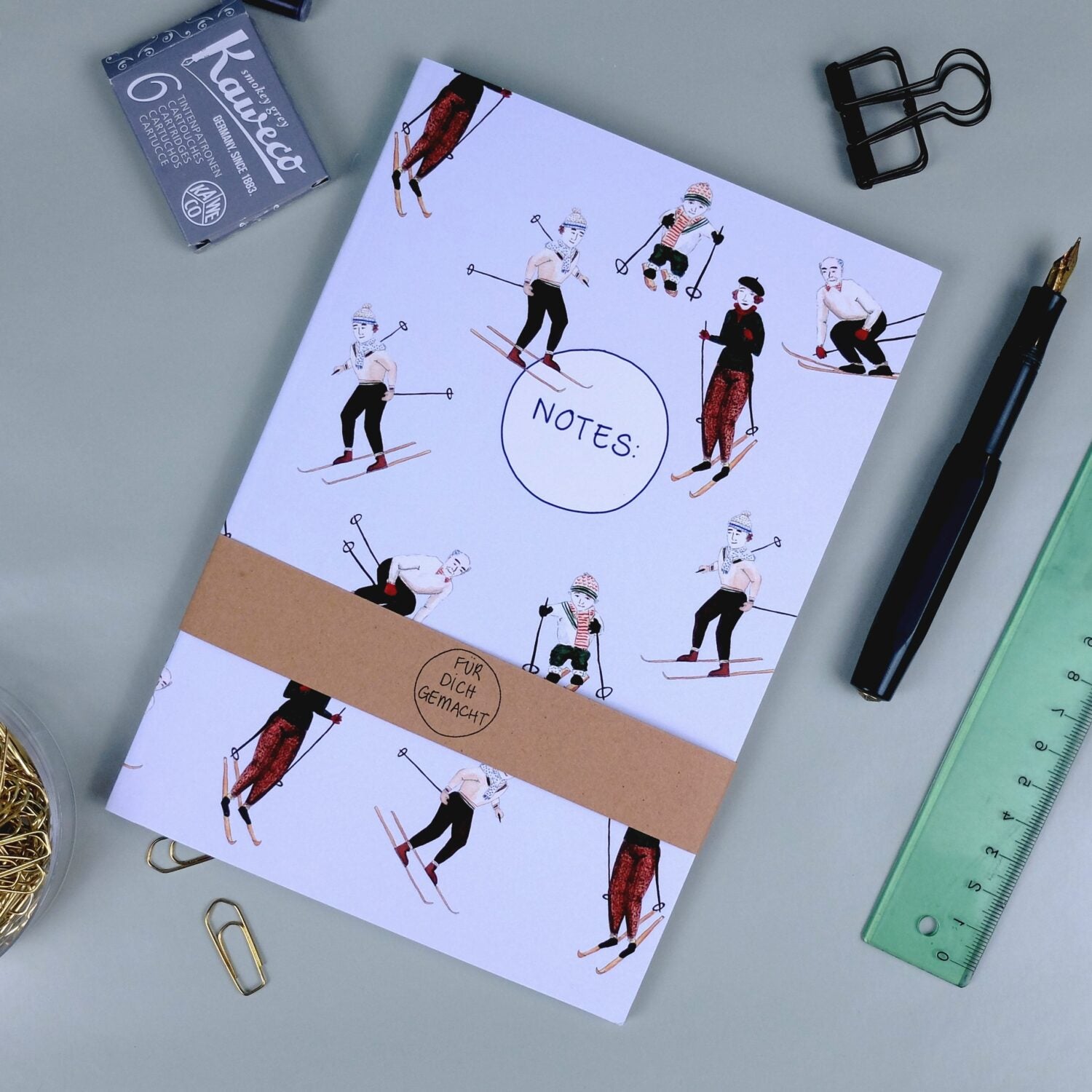 Notizbuch mit Skifahrerillustrationen, umgeben von Büroartikeln wie Stift, Lineal, Klammern und Radiergummi.