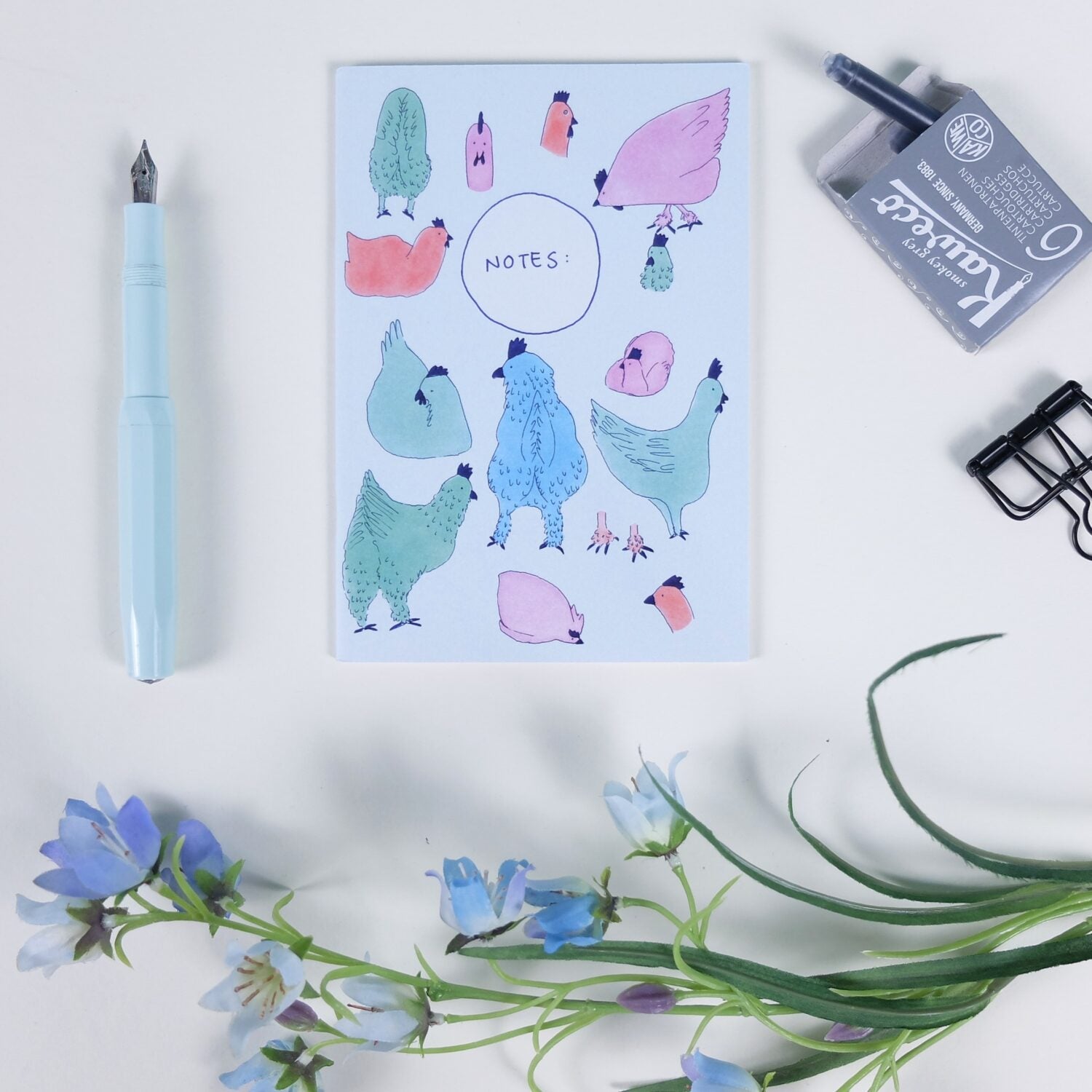 Notizbuch mit bunten Hühnerbildern, neben einem blauen Füller, Tintenpatrone, Klammer und Blumen auf weißem Hintergrund.